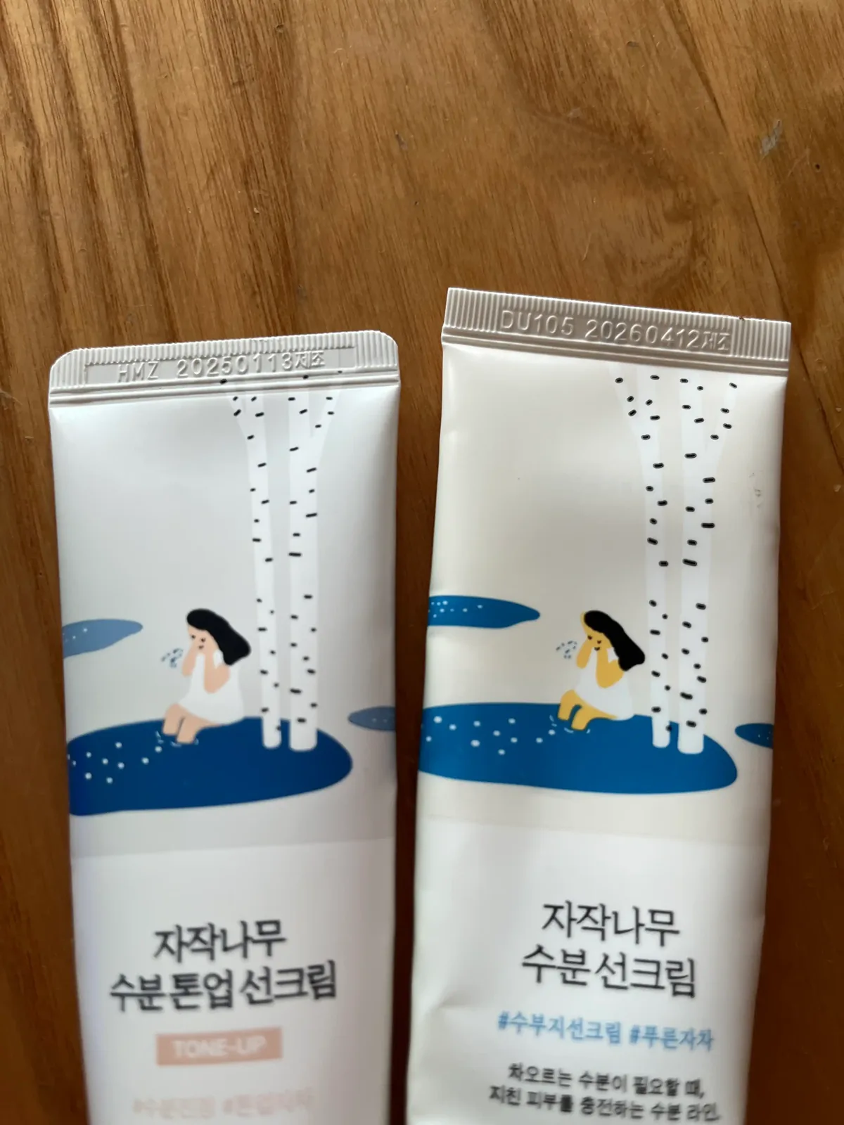 자작나무와 물가에 앉은 인물 일러스트가 그려진 분홍색과 남색 선크림 튜브 앞면 윗부분의 제조 코드